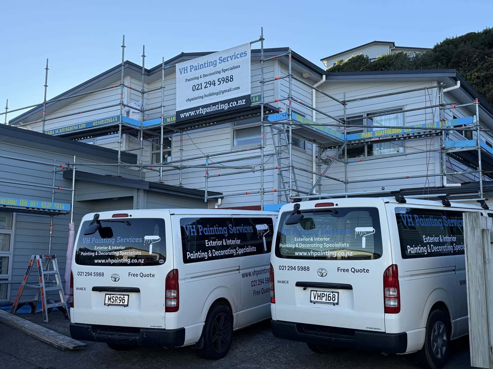 Two VH Painting vans outside a house under scaffolding, with a VH Painting banner on the scaffold